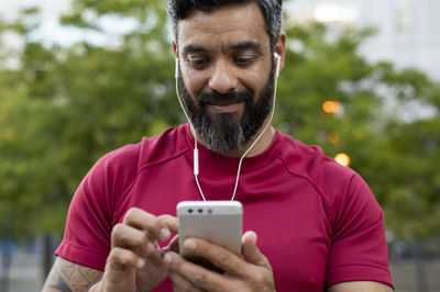 Low angle view of smiling confident man using smart phone while standing against trees in park