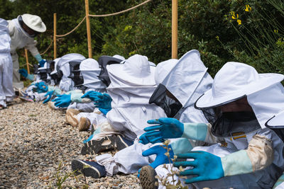 Side view of group of anonymous children and beekeepers in safety costumes sitting on ground in apiary