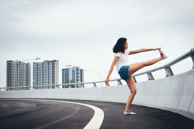 Full length side view of woman exercising by railing on bridge in city against sky
