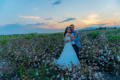 Portrait of bride and groom standing on field against sky during sunset