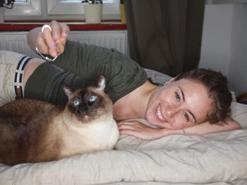 Portrait of happy woman lying by siamese cat on bed at home