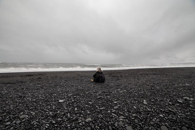 Rear view of woman sitting on pebbles at beach against sky