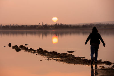 Rear view of silhouette man standing by lake against sky during sunset