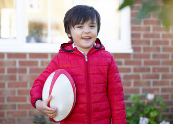 Portrait of smiling boy holding american football while standing against brick wall