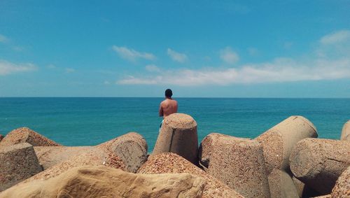 Rear view of shirtless man on tetrapods at sea shore against sky