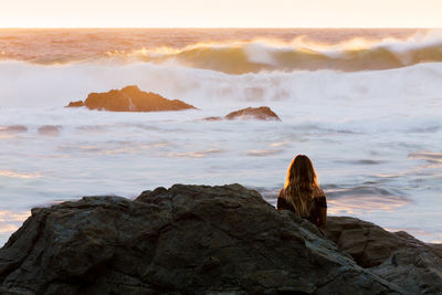 Rear view of woman sitting on rock by sea against sky