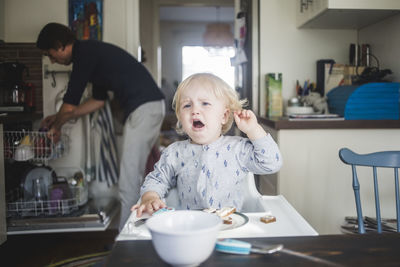 Crying male toddler looking away while sitting on high chair in kitchen at apartment