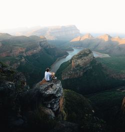 Man sitting on rock looking at mountain against sky