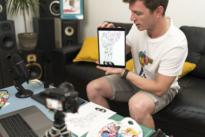 Male artist showing digital tablet while live streaming on laptop at home