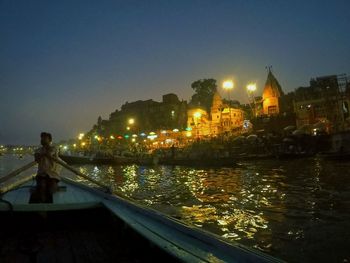 Illuminated city by river against sky at dusk