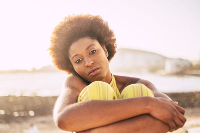 Portrait of young woman hugging knees while sitting against sky during sunset