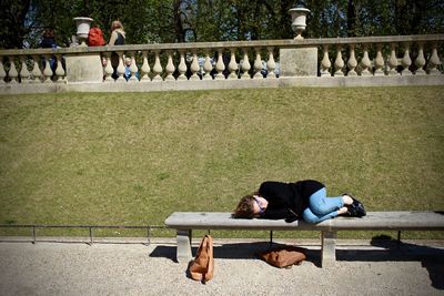 Young woman lying on bench against retaining wall in park during sunny day