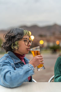 Delighted young woman sitting at table in veranda of pub in city of cusco and drinking glass of beer and looking away