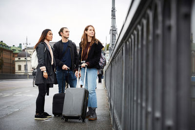 Full length of male and female friends with luggage standing on bridge in city