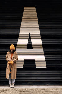 Full body of female with handbag in coat and hat standing near building with closed metal shutter on street of city