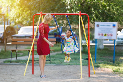 Side view of woman looking at daughter playing on swing at playground