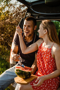 Millennial couple sitting on open trunk and eating watermelon. happy young couple having break at