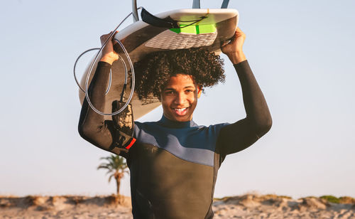 Happy mixed race surfer boy balancing a surfboard on his directly at the camera and smiling.