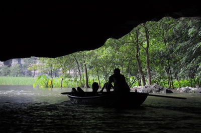 Rear view of silhouette men in boat sailing on river below cave