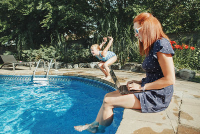 Side view of woman using laptop while girl jumping in swimming pool