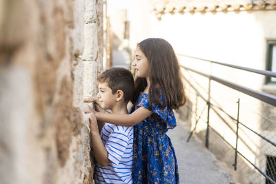 Side view of siblings looking through old window while standing outdoors