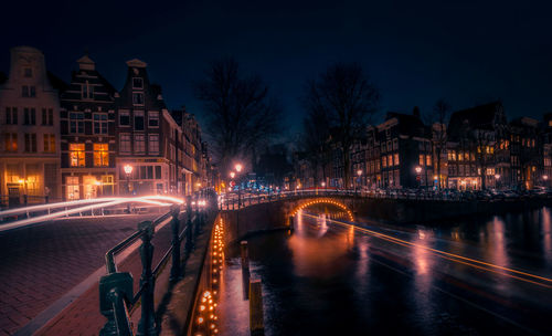 Illuminated bridge over river by buildings against sky at night