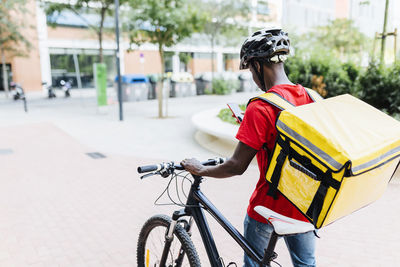 Delivery man using smart phone while wheeling bicycle on footpath