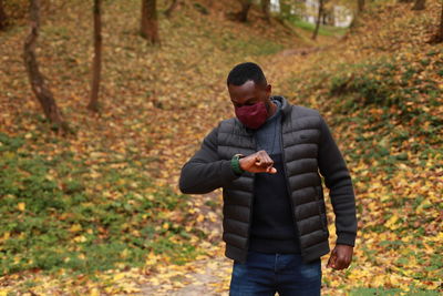 Black man in fabric anti-covid facial mask checking time and standing in park during autumn