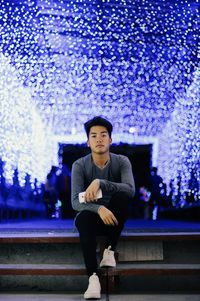 Portrait of young man sitting against illuminated christmas decoration at night