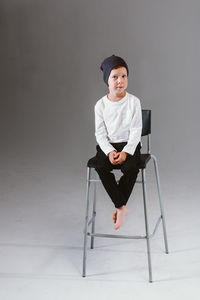 A boy in a white t-shirt and cap sits on a bar stool, on a white background. 