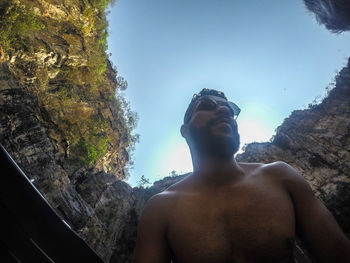 Low angle view of shirtless man looking at rock formation against sky