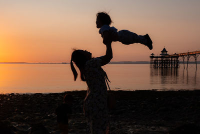 Silhouette woman standing on beach with toddler against sky during sunset