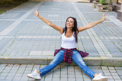 Happy girl in white t-shirt, blue jeans and white sneakers sits on sidewalk