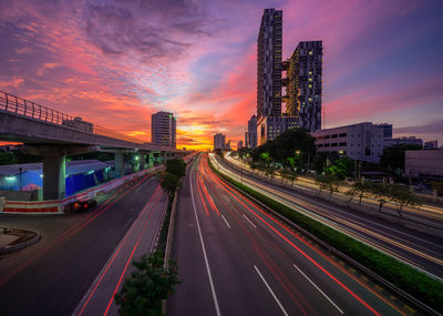 Light trails on road amidst buildings against sky during sunset