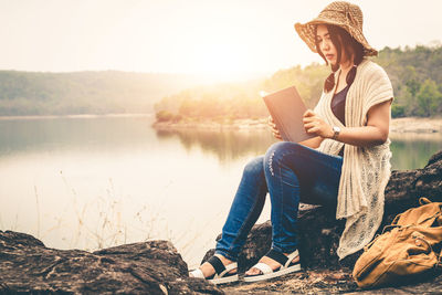 Young woman sitting on book by lake against sky