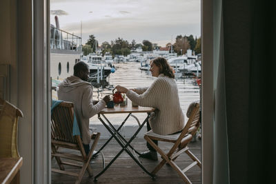 Rear view of couple having tea while sitting on chair at porch by marina