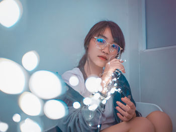 Portrait of young woman with illuminated string lights sitting at home