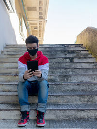 Young caucasian man dressing casual watching a tablet, while sitting on stairs, with a  face mask
