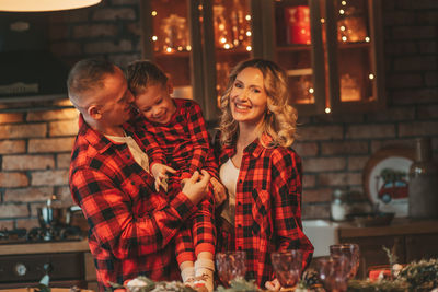 Smile active parents with small son in red checkered sleepwears waiting santa indoor.