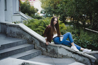 Young woman holding coffee cup while sitting on retaining wall