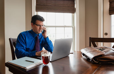 Man on cellphone working from home using a computer at a dining table.