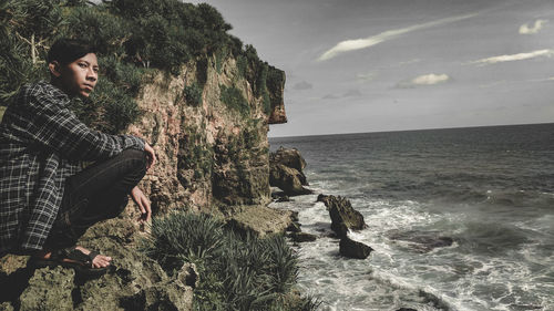 Young man on rock by sea against sky