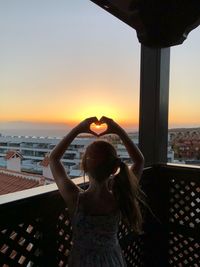 Rear view of girl making heart shape while standing at balcony during sunny day