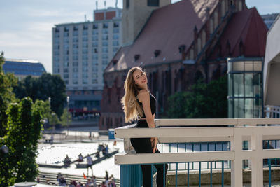 Side view portrait of beautiful young woman standing on balcony against buildings in city