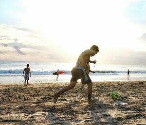 Young men playing soccer on sand at beach against sky during sunset
