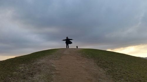 Low angle view of man with arms outstretched standing on hill against cloudy sky