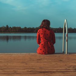 Rear view of woman sitting on pier over lake against sky