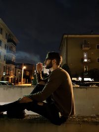 Boy smoking while sitting on street at night against sky