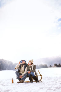 Back view of senior couple sitting side by side on sledge in snow-covered landscape drinking hot beverages