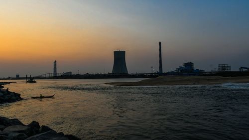 Silhouette factory by sea against sky during sunset